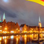 Night view of Bremen cityscape with historic buildings illuminated along the river, reflecting colorful lights on the water, with a German flag graphic in the corner.